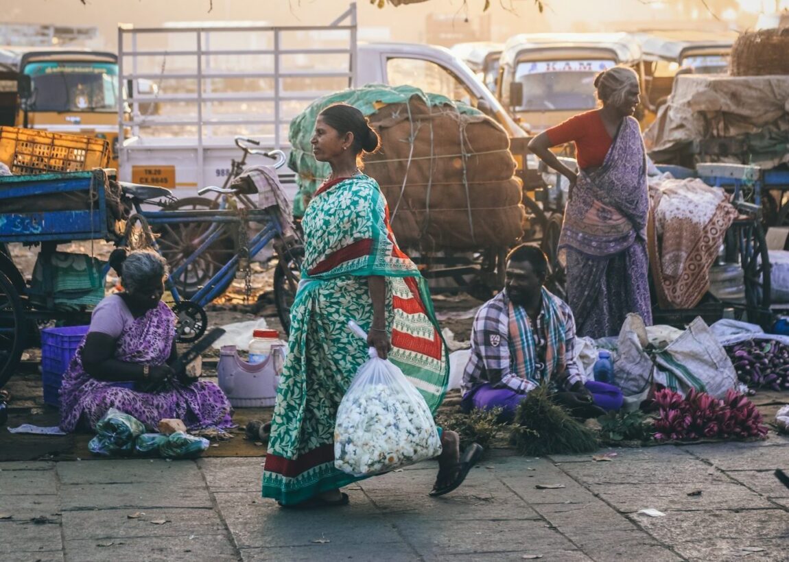 A bustling outdoor market scene, featuring a woman in a green sari walking with a bag in hand, while other vendors sit and stand among fresh produce, bicycles, and auto-rickshaws in the background.
